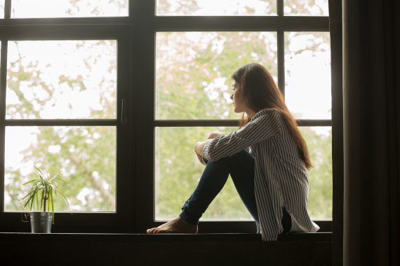 Thoughtful,Girl,Sitting,On,Sill,Embracing,Knees,Looking,At,Window,